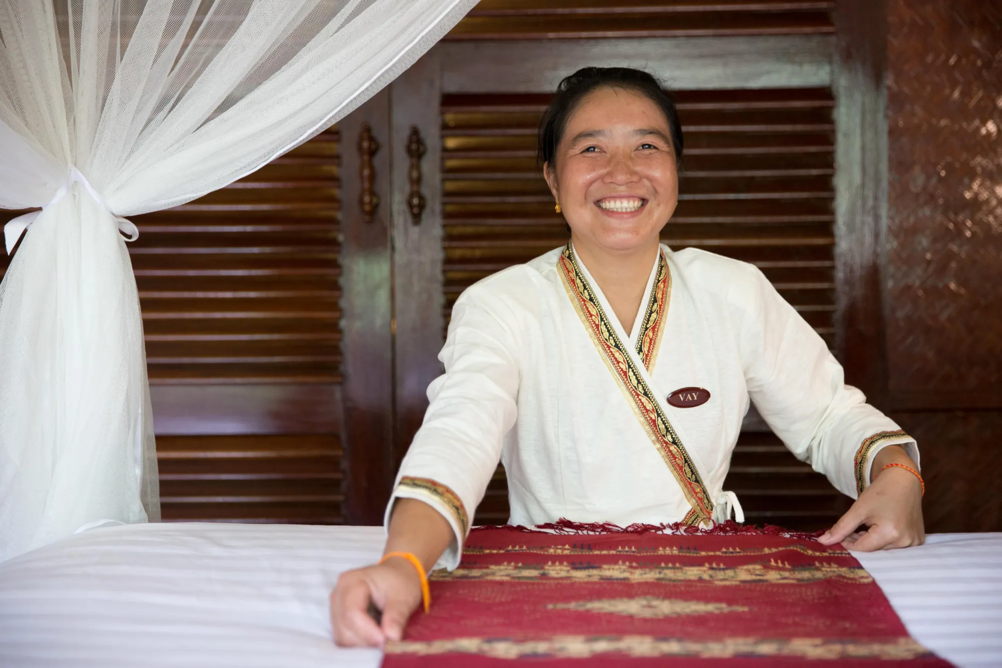 Une membre du personnel du lodge souriante, en tenue traditionnelle, préparant un lit avec un jeté en coton tissé