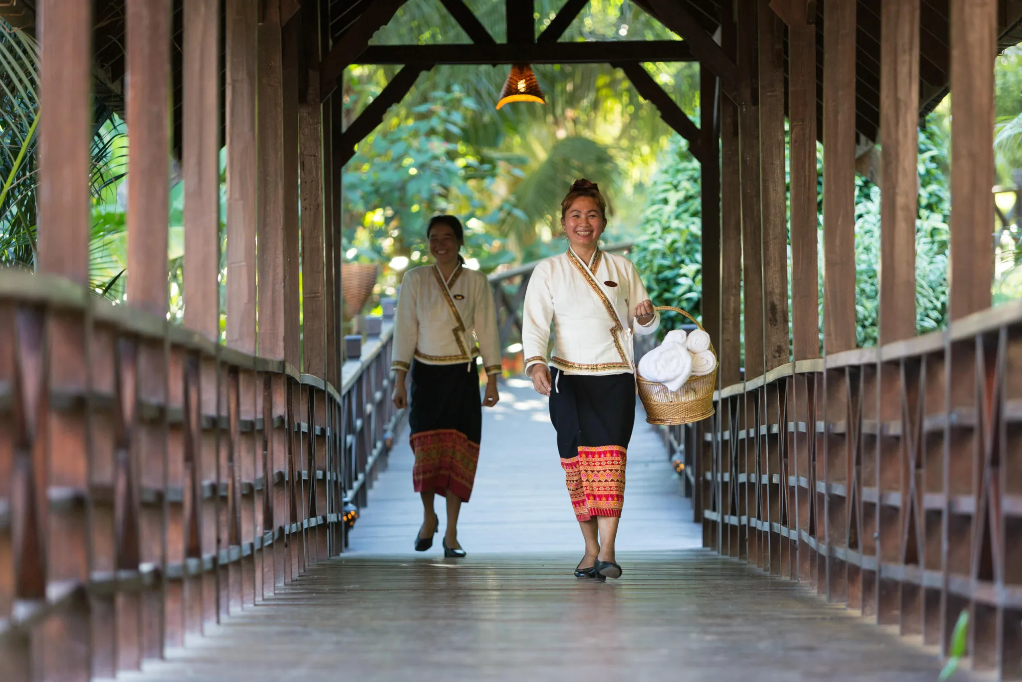 Deux membres du personnel en tenue traditionnelle lao traversant une passerelle en bois couverte, l'une portant un panier de serviettes propres