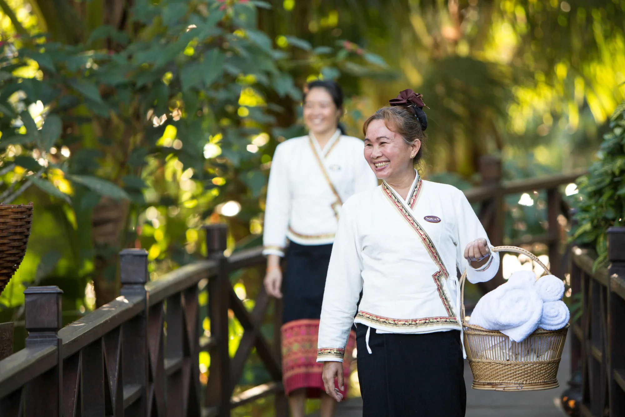 Des membres du personnel en tenue traditionnelle souriant en traversant les jardins avec un panier de serviettes