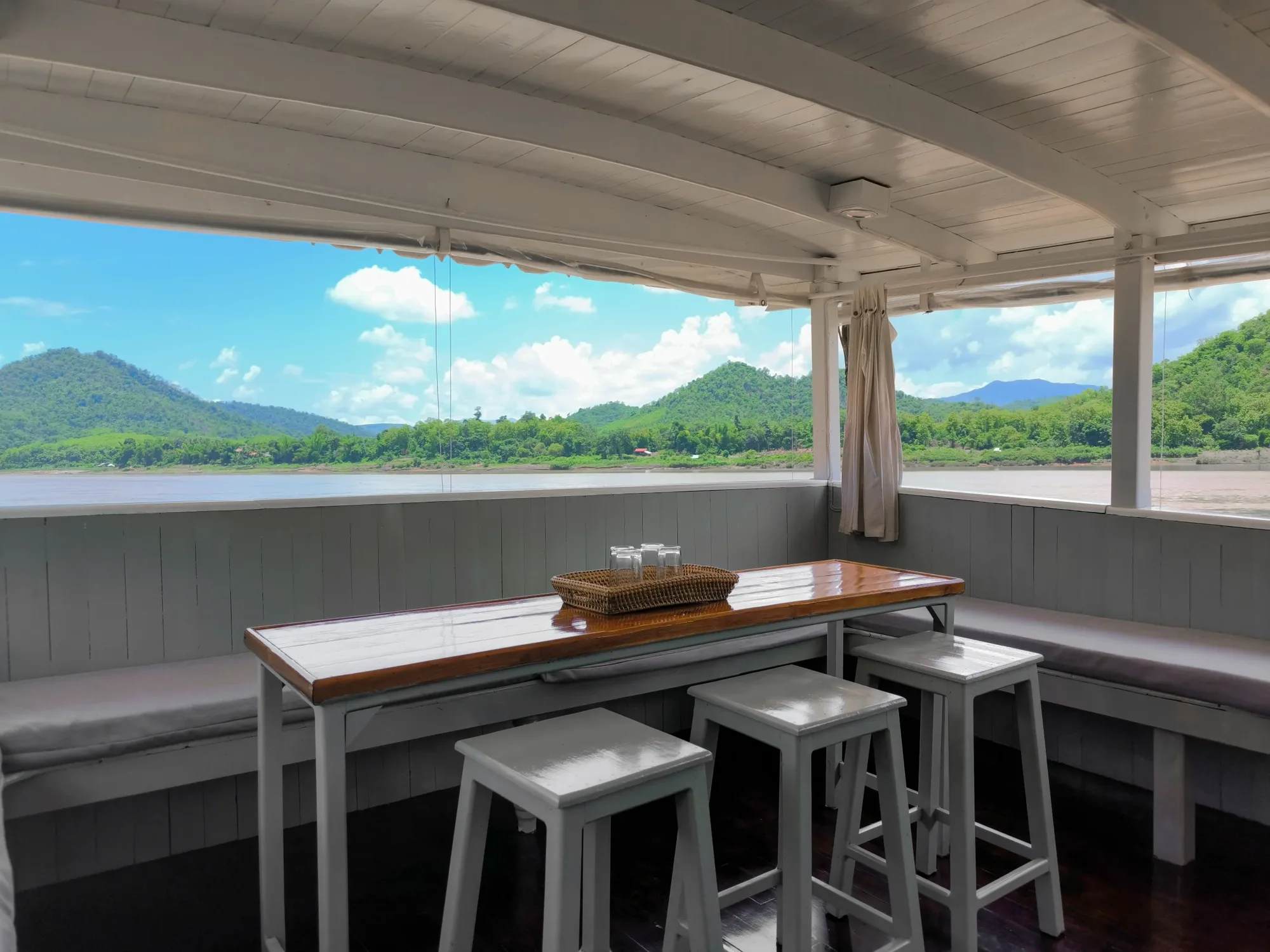 A wooden table and stools on the open deck of the Mekong Cruises boat, green hills across the river