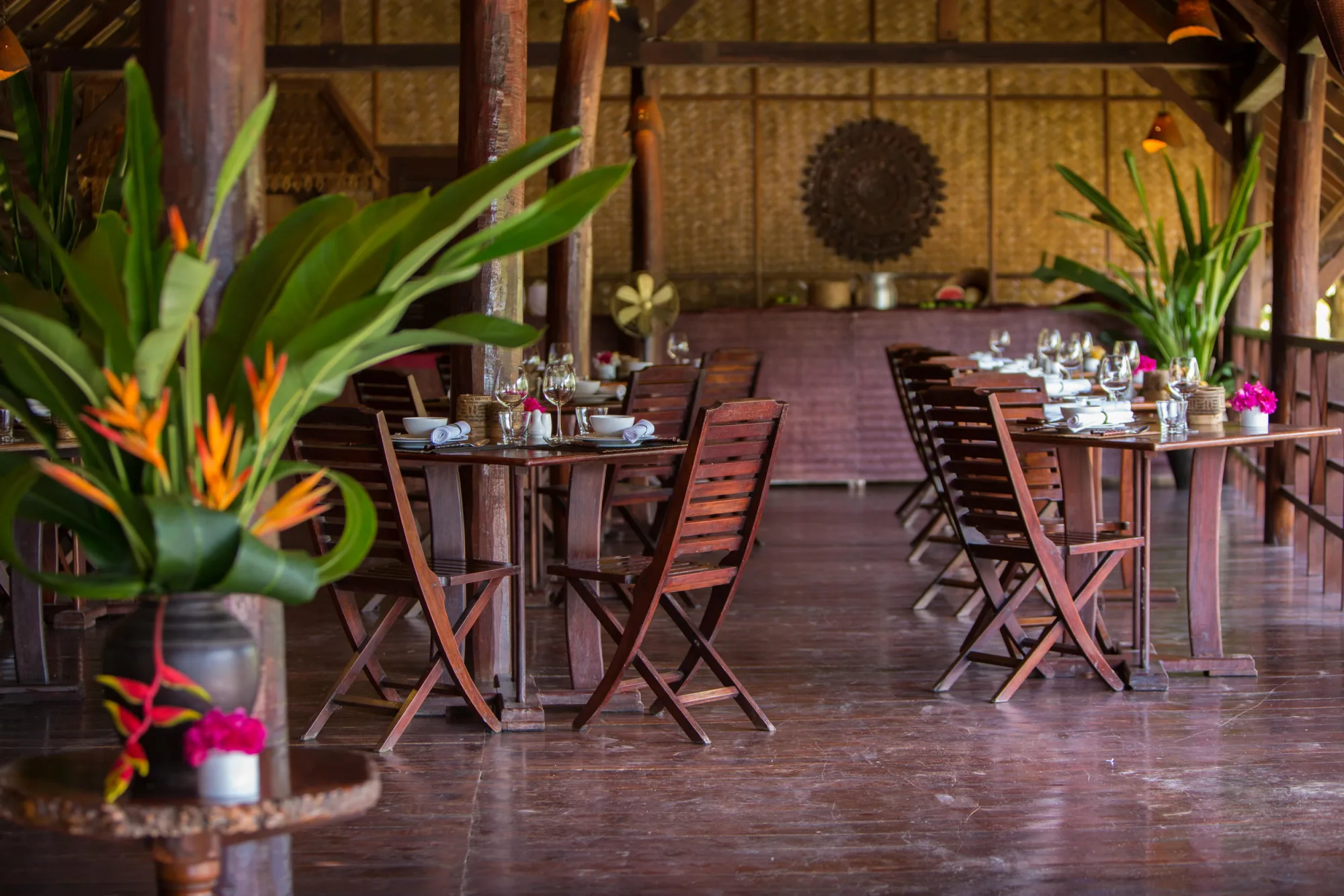 Salle à manger au bord du fleuve sous un haut toit de chaume, tables dressées avec des compositions florales tropicales