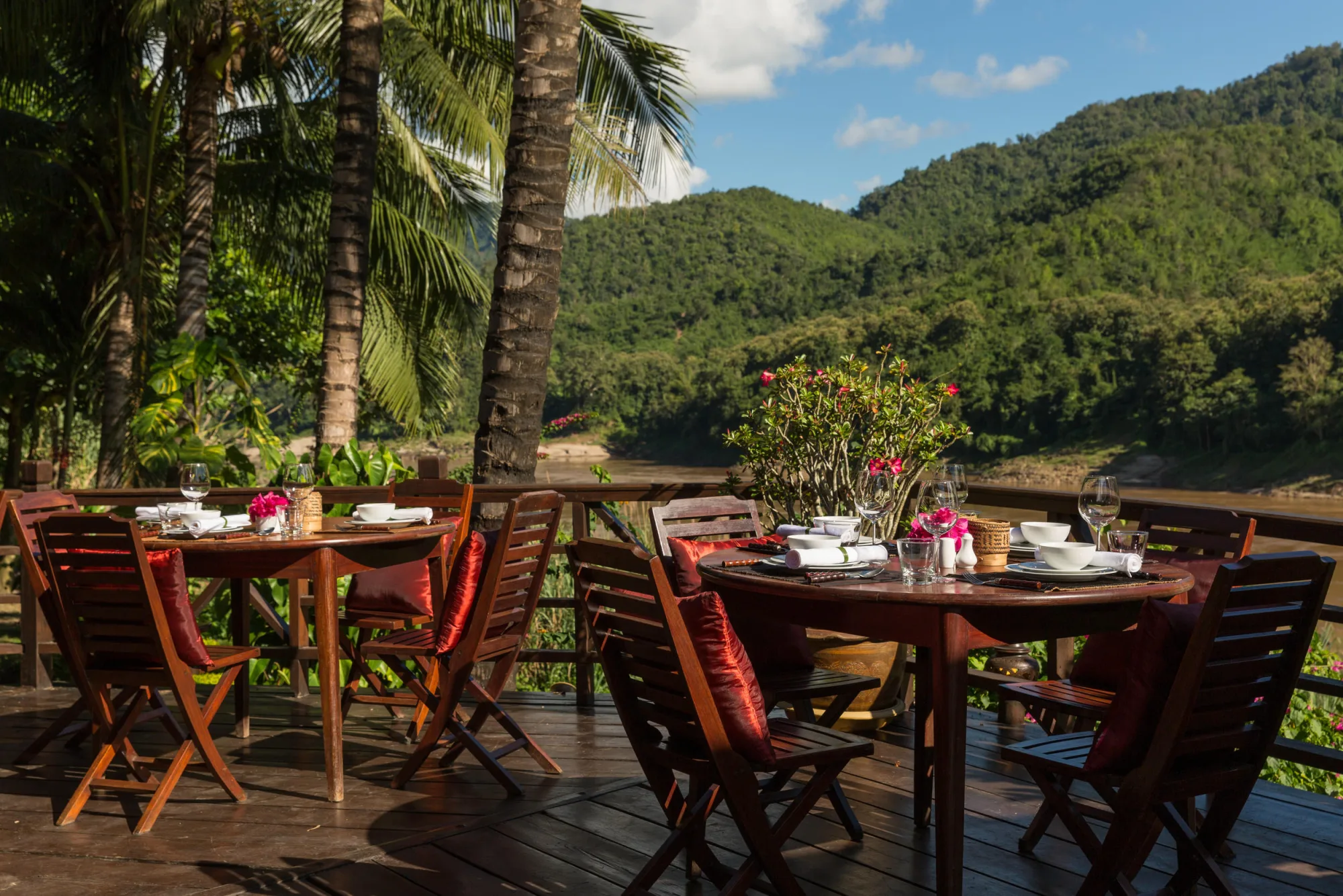 Tables rondes dressées pour le repas sur la terrasse parmi les palmiers, avec le Mékong et les collines boisées en arrière-plan