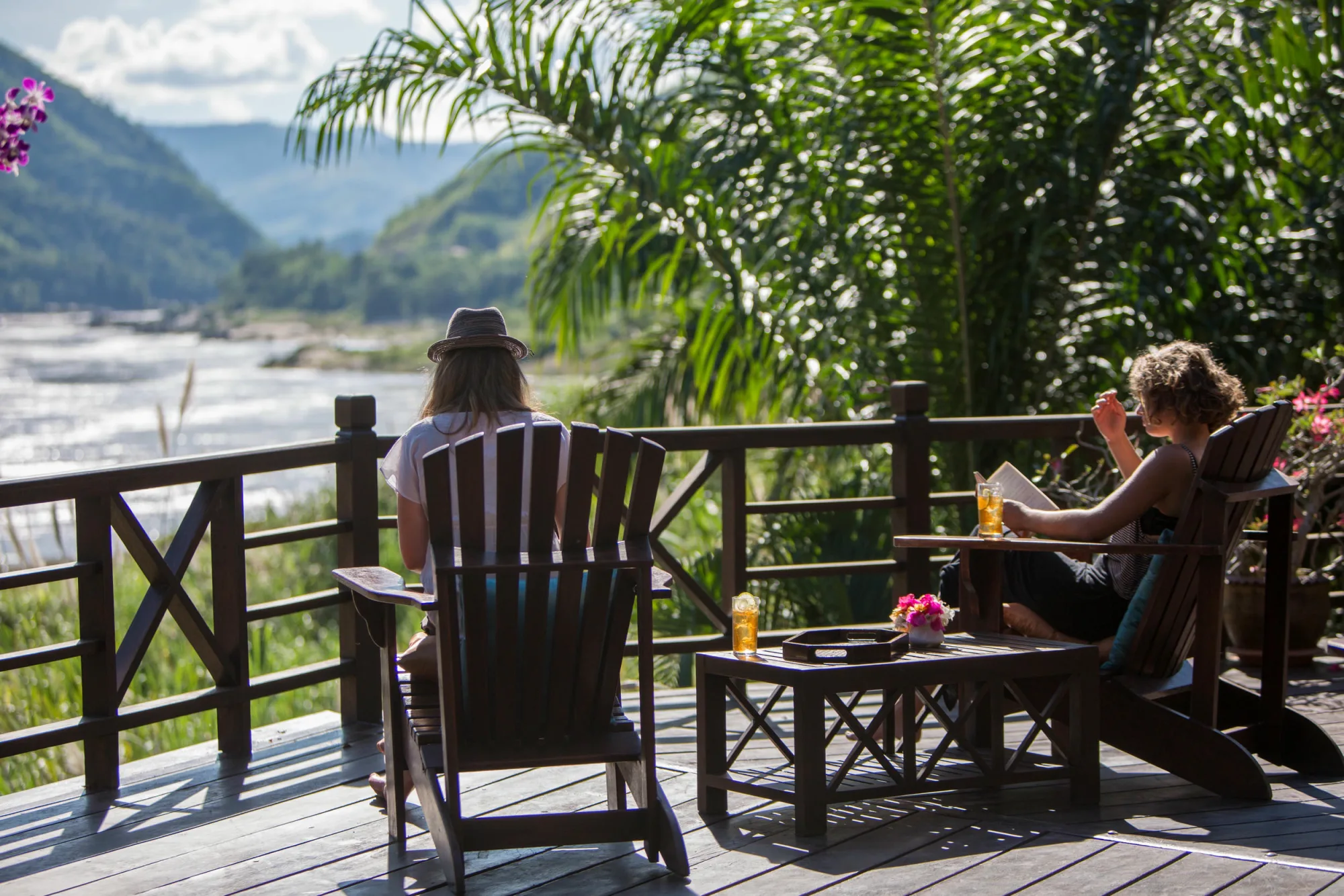 Des clients installés dans des fauteuils en bois sur la terrasse au-dessus du Mékong, avec des boissons et un livre
