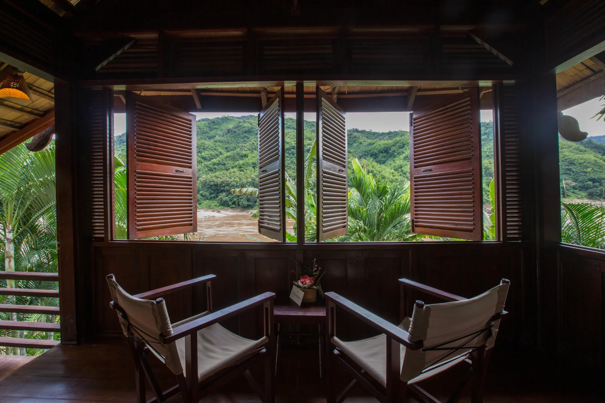 Two armchairs by open wooden shutters looking out over the Mekong from inside a bungalow