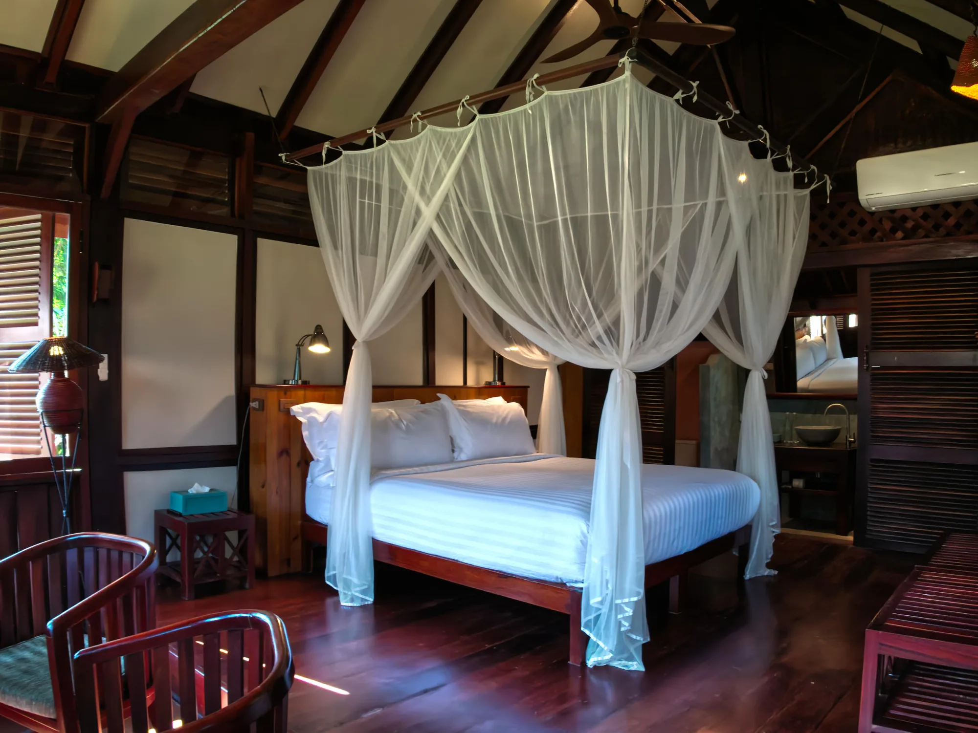 King bed under a white mosquito net in a teak bungalow with a high timber ceiling
