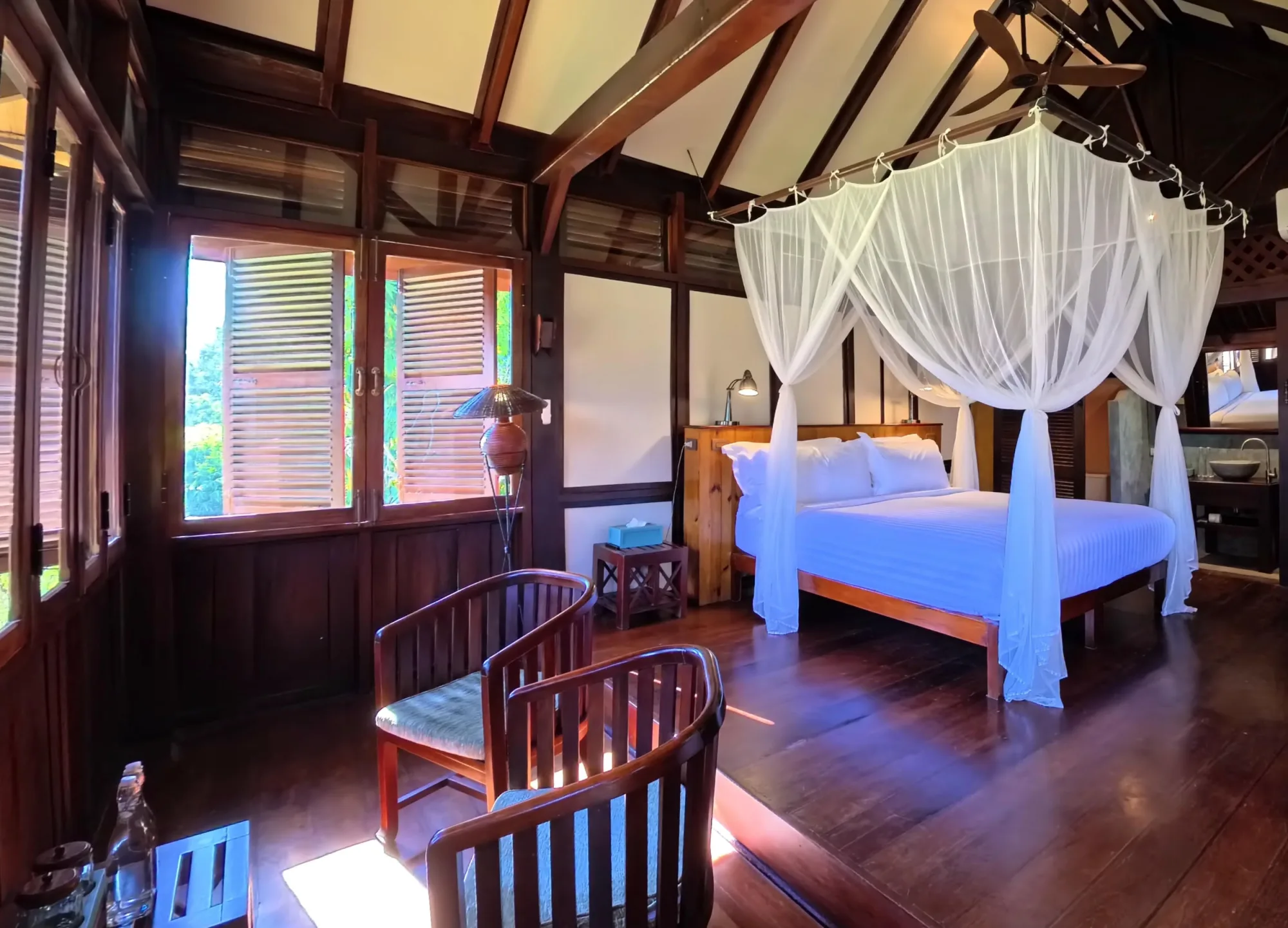Intérieur d'un bungalow avec lit king, moustiquaire, fauteuils en bois et fenêtres à volets