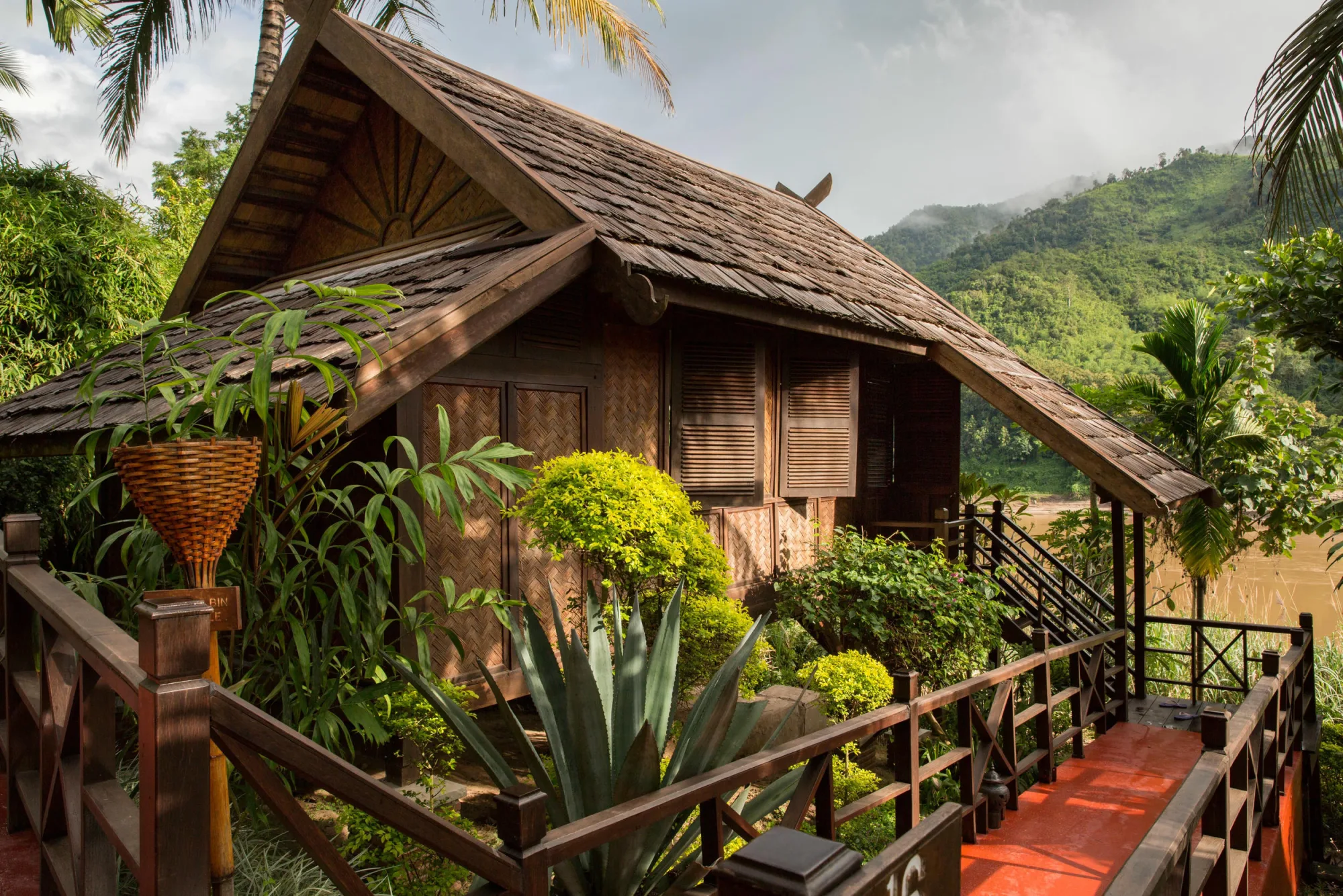 Front of a bungalow with a wooden railing and tropical garden, mountains behind