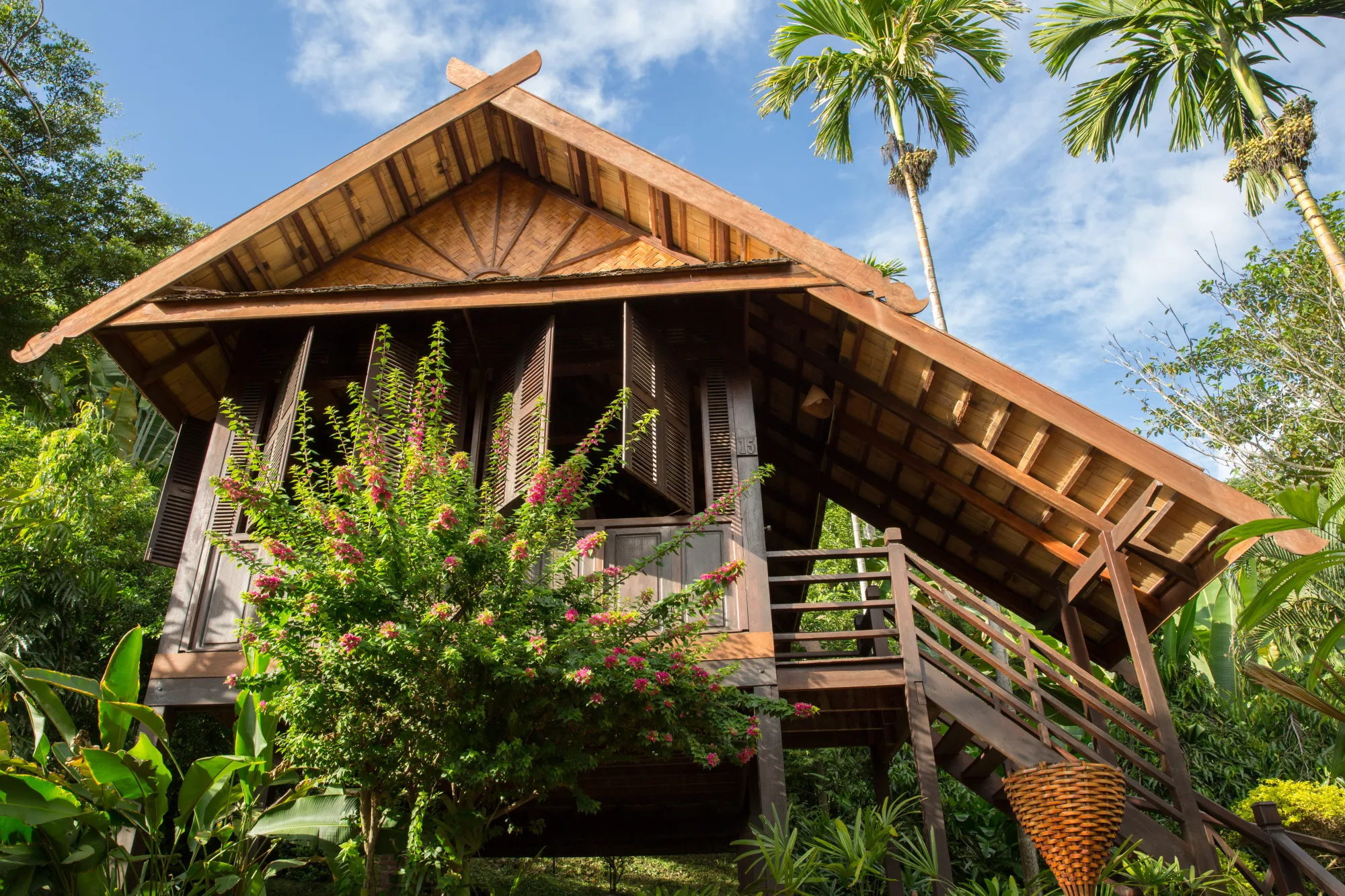 Bungalow en teck au toit pointu, volets en bois ouverts et escalier, entouré de bougainvilliers et de palmiers